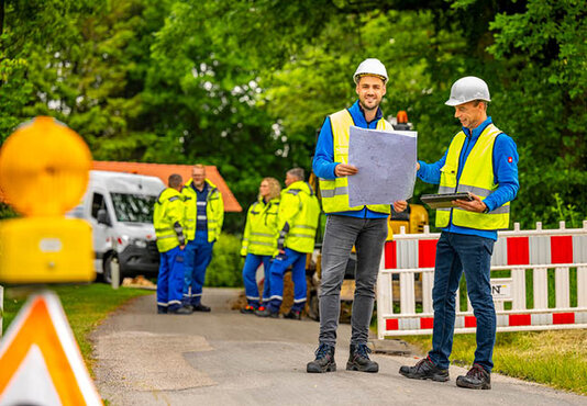 Zwei Techniker stehen vor einer Baustelle und haben einen Plan in der Hand